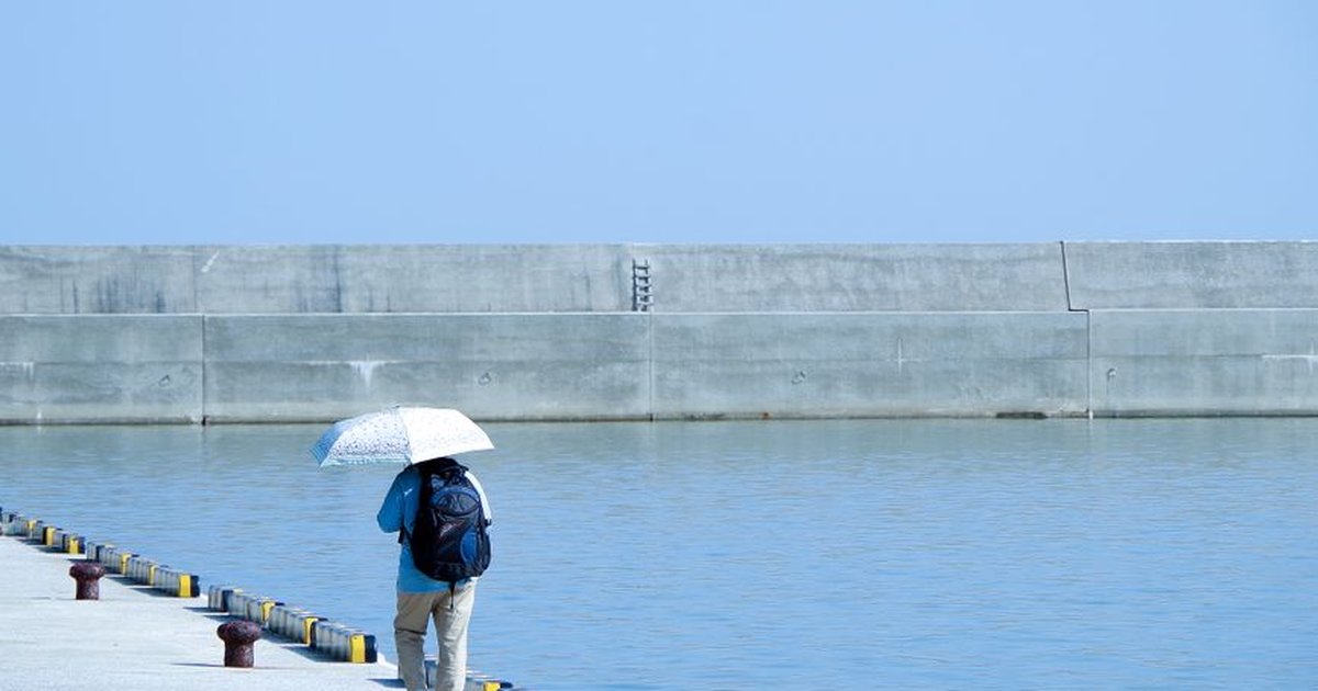 A solitary figure with umbrella walks along a sunny pier beside calm blue waters.
