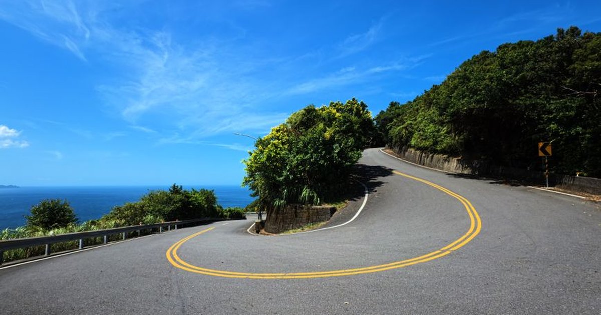 A picturesque curved road with double yellow lines, surrounded by lush greenery and overlooking the ocean in New Taipei City, Taiwan.