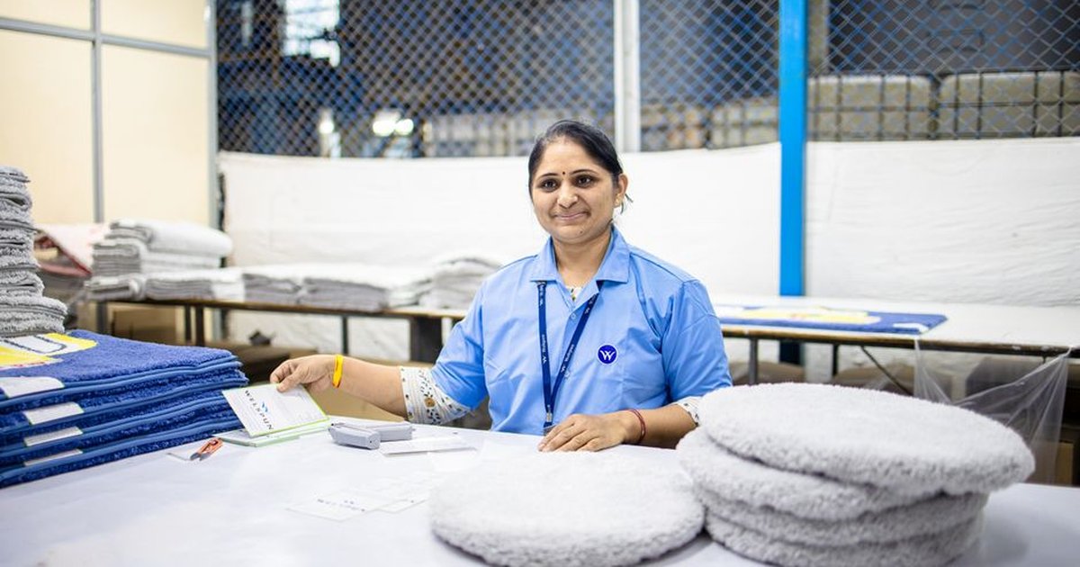 Female textile worker smiling in a factory surrounded by fabrics and packaged products.