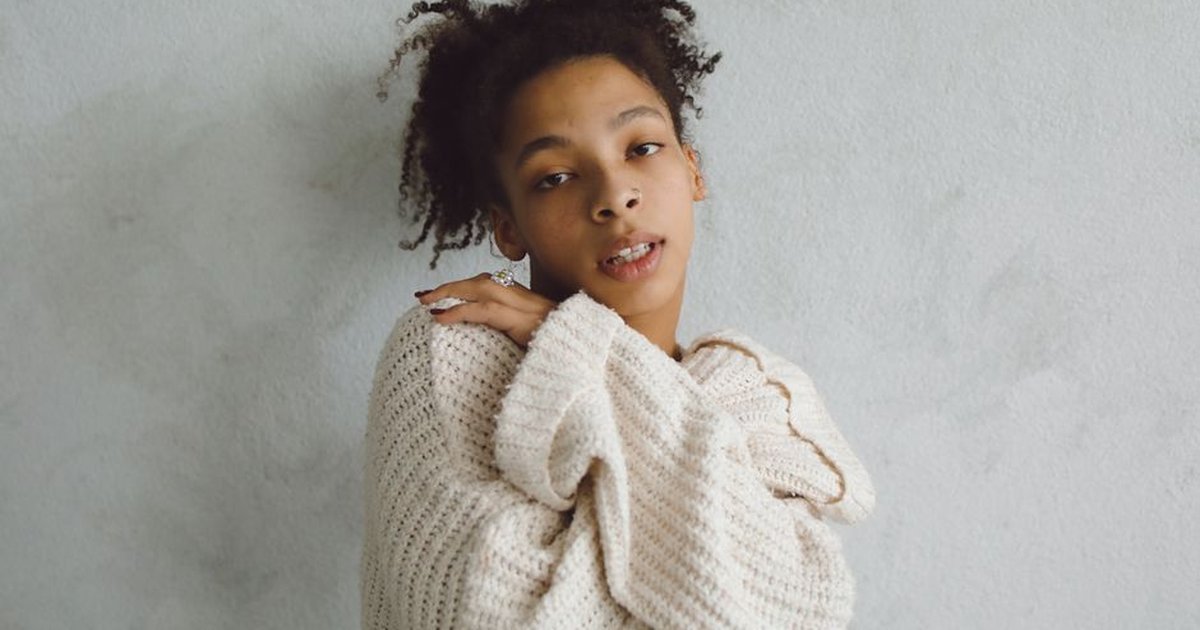 A woman poses with arms crossed in a cozy knit sweater against a white wall.