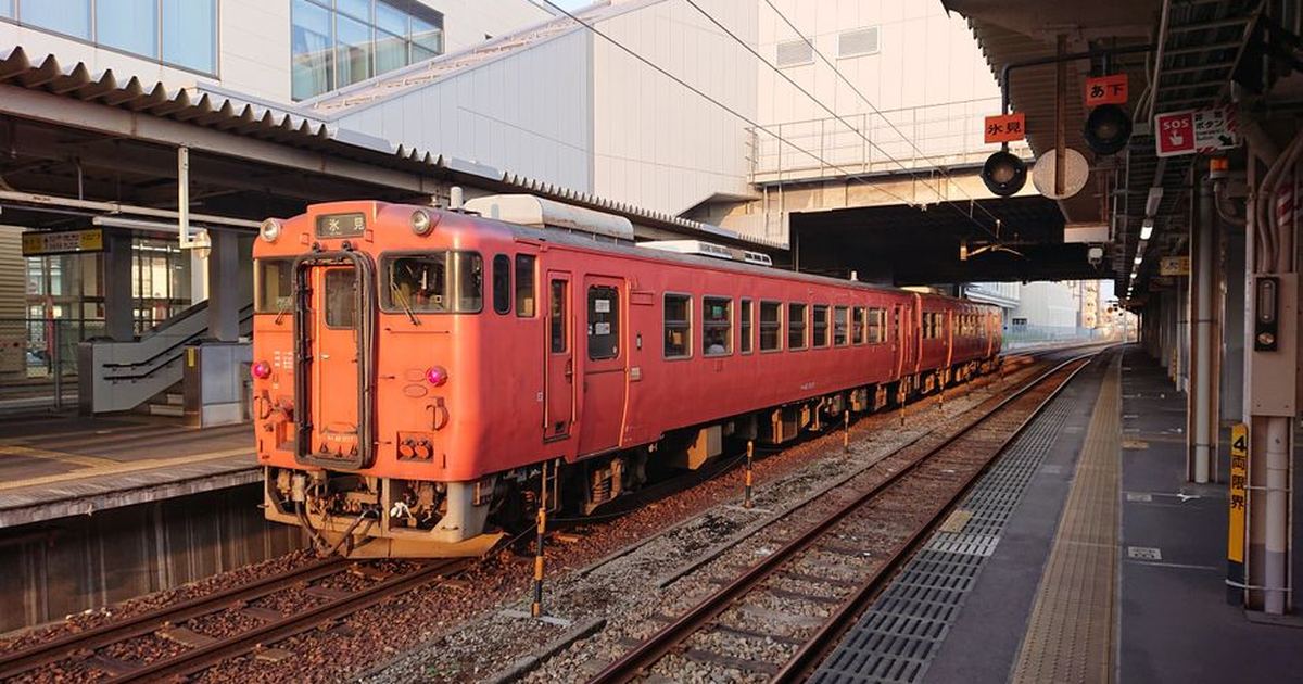 A red train at Toyama Station platform, bustling with urban architecture under daylight.