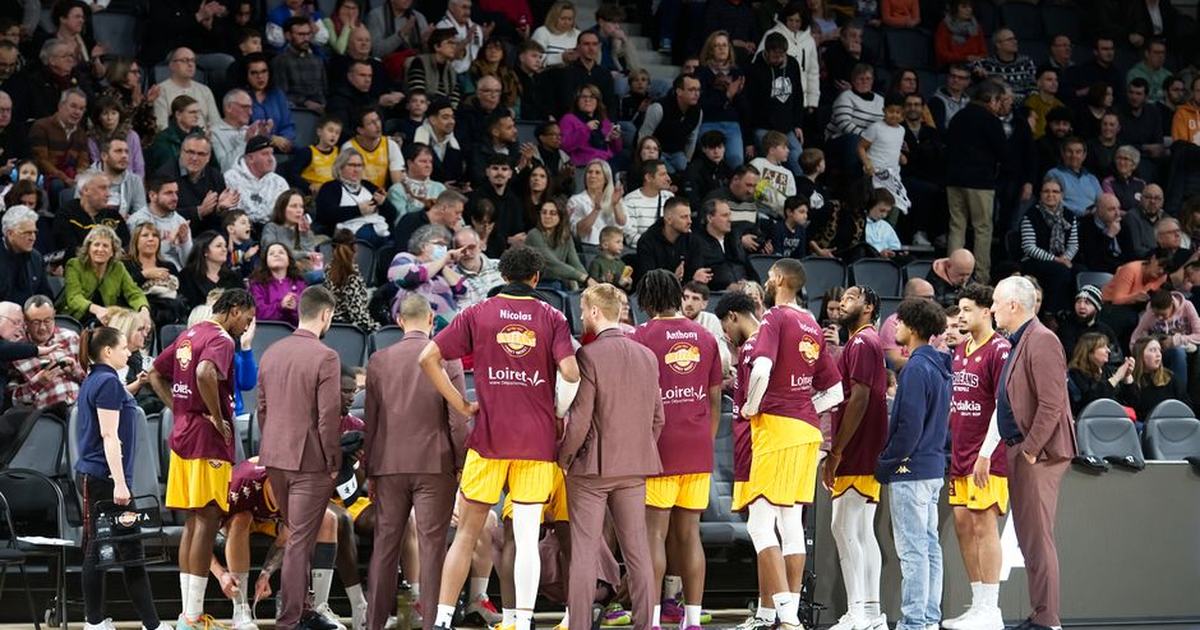 Basketball team huddles on court during a game timeout with audience in background.