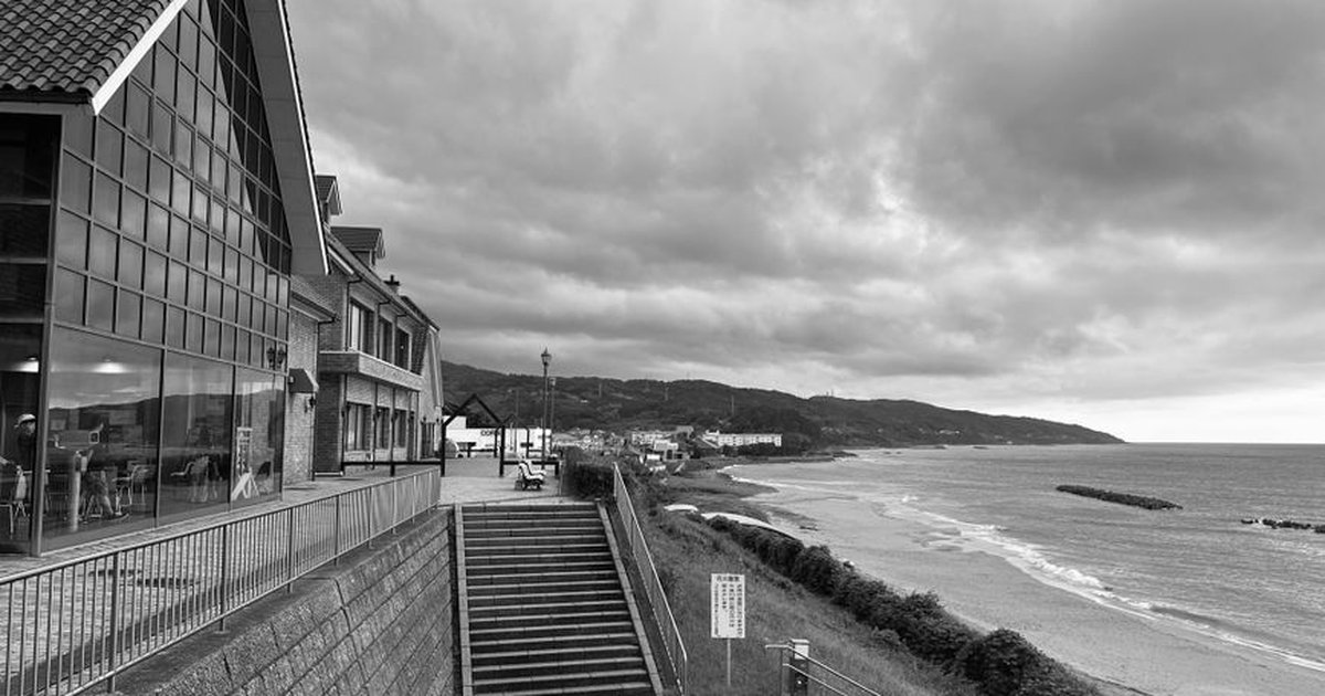 A black and white photograph of a coastal view featuring a modern building and a dramatic cloudy sky.